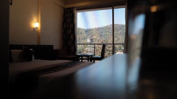 A hotel room with a bed, a small table, and a view of a mountainous landscape through a large window. - Royal Hills Vasai - Thane (Vasai)