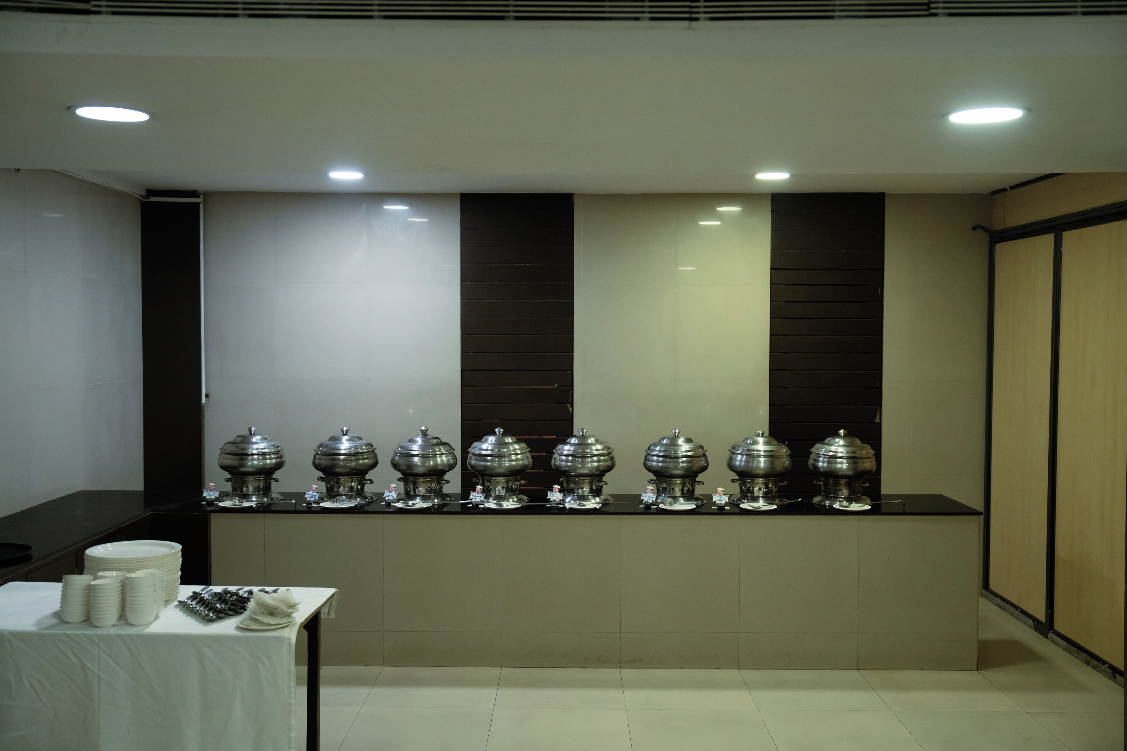 Montaje de bufé con varios dispensadores de comida y una mesa con platos y cubiertos apilados.