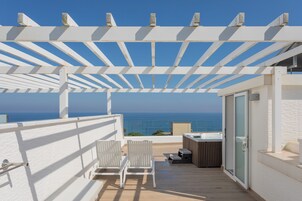 Un balcón con una pérgola blanca, dos sillas blancas, un jacuzzi y vista al océano.