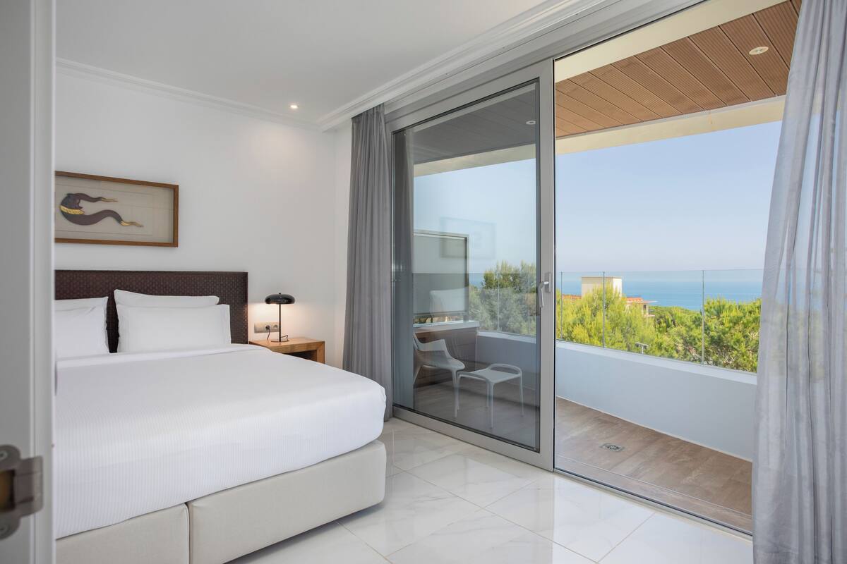 A modern hotel room with a large bed, a desk, and a balcony with a view of the sea.