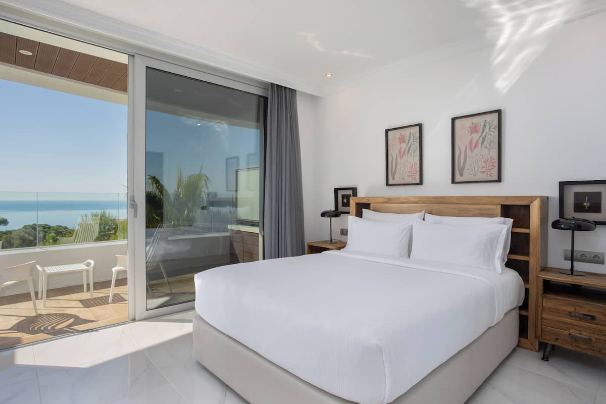 A modern bedroom with a large bed, a glass door leading to a balcony, and two framed botanical prints on the wall.