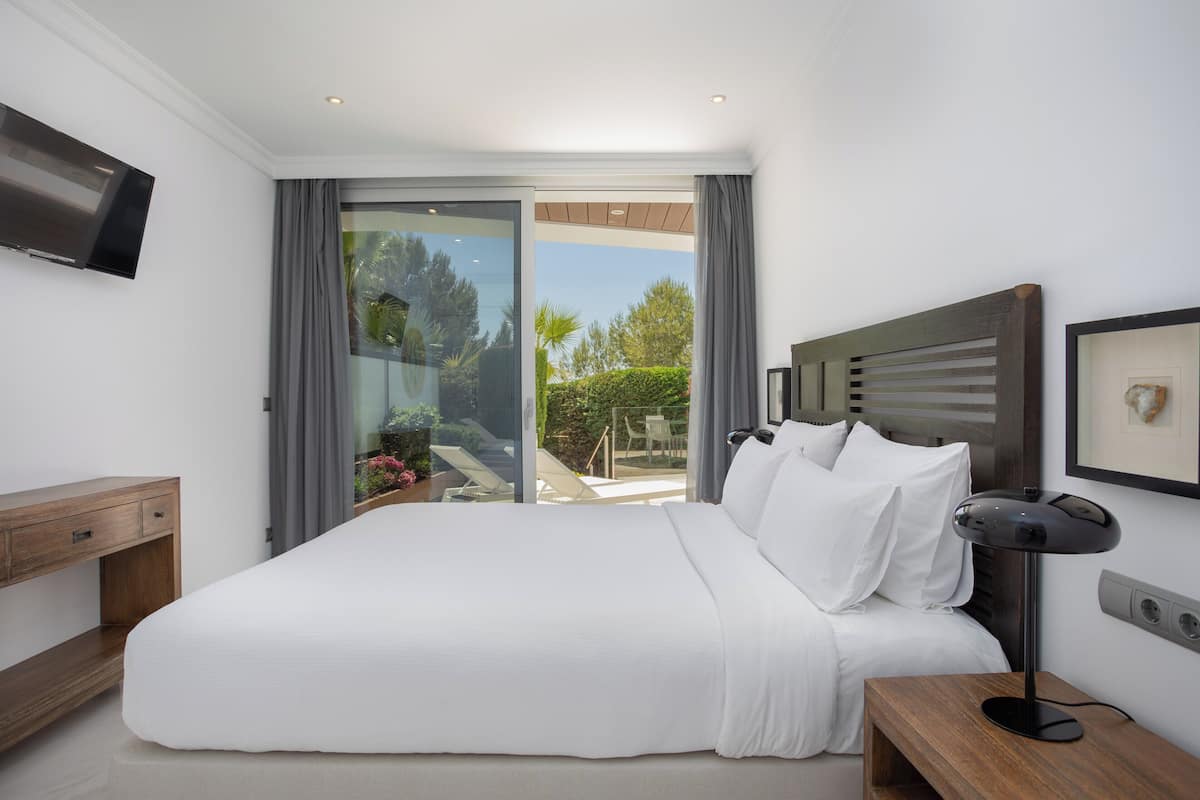 A bedroom with a large bed, a TV, a bedside table, and a view of the outdoors.