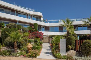 Un hotel moderno con estructura de varios niveles, un jardín con diversas plantas y una fuente de piedra, bajo un cielo azul despejado.
