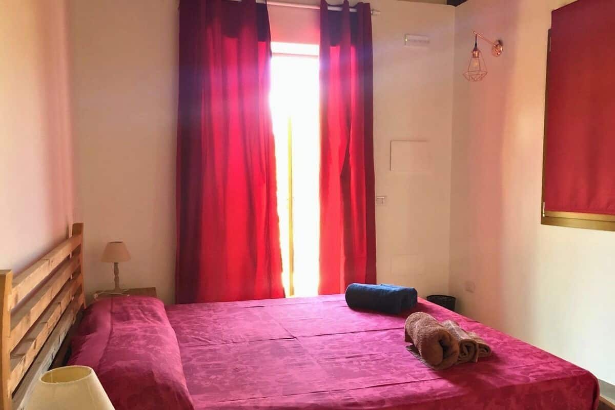 A small, neatly made bed with a pink bedspread, a rolled towel, and a lamp on a bedside table.