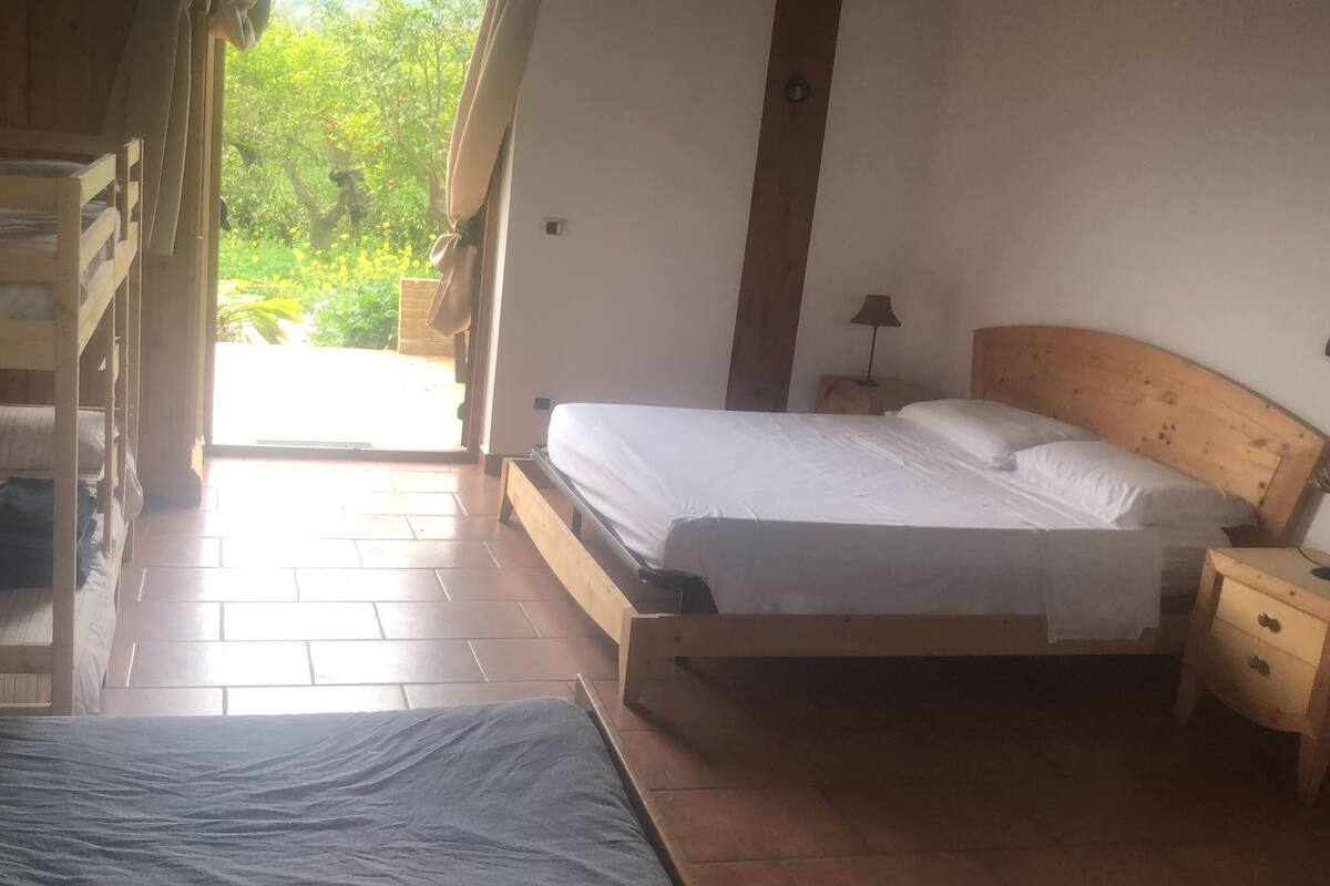 A room with two wooden beds, one with a blue bedspread and the other with white bedding. There is a wooden nightstand with a lamp, and a window with curtains letting in natural light.