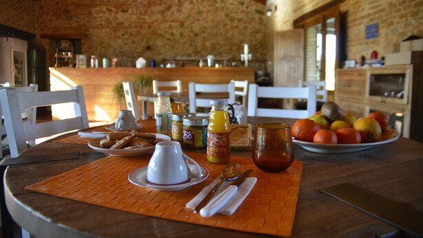Een rustieke eetruimte met een stenen muur, houten plafond en een tafel gedekt met ontbijtartikelen zoals fruit, brood en drankjes.