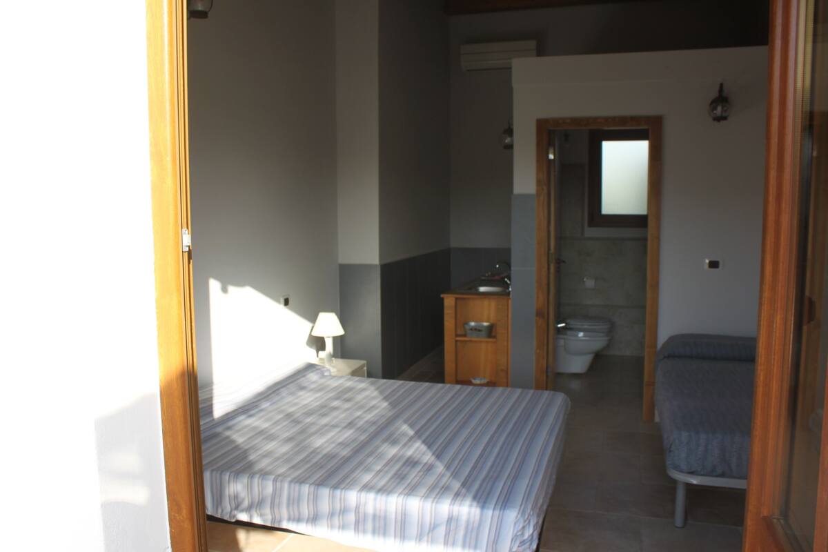 A small, well-lit bedroom with a bed, a bedside table with a lamp, a wooden cabinet, a bathroom with a toilet and sink, and a mirror on the wall.