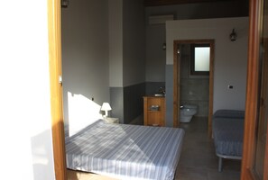 Une petite chambre bien éclairée, avec un lit, une table de chevet dotée d’une lampe, une armoire en bois, une salle de bain avec des toilettes et un lavabo, et un miroir au mur.