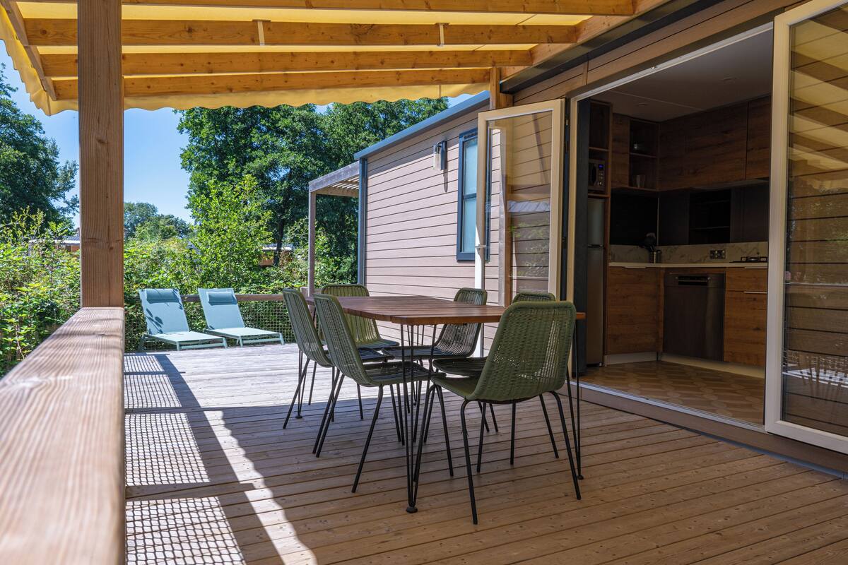 Eine Holzterrasse mit Tisch und Stühlen, eine Schiebetür zu einer Küche und Blick ins Grüne.