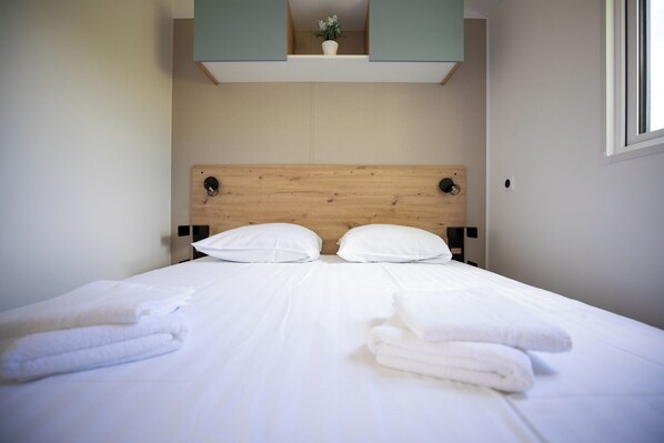 A neatly made bed with white linens and towels, a wooden headboard, and a small potted plant on the nightstand. - RCN Camping La ferme du Latois (Coëx)
