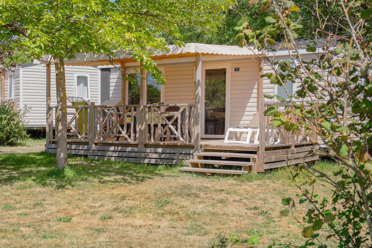 Ein Mobilheim mit Veranda und kleiner Terrasse, umgeben von Gras und Bäumen.