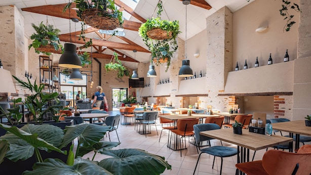A spacious cafe with high ceilings, exposed brick walls, and hanging plants. Tables and chairs are arranged for dining. - RCN Camping La ferme du Latois (Coëx)