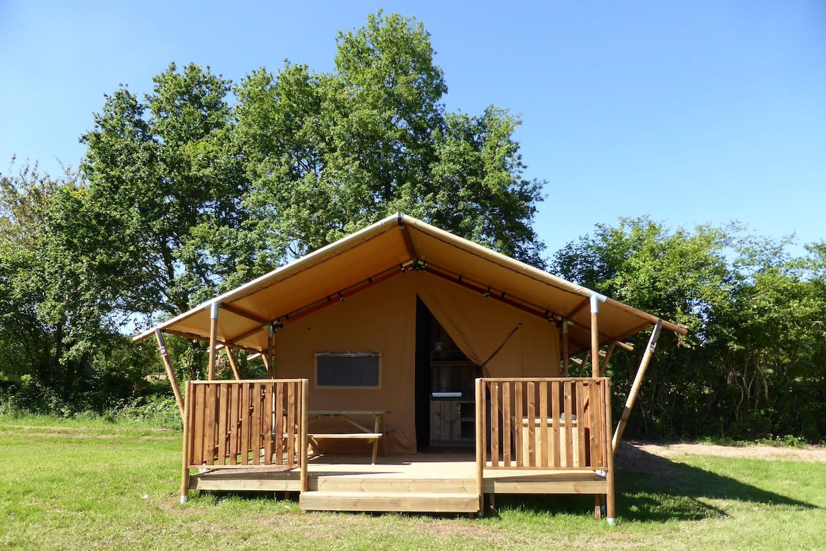 Ein einstöckiges, offenes Holzgebäude mit Veranda und einer Bank, umgeben von Gras und Bäumen.