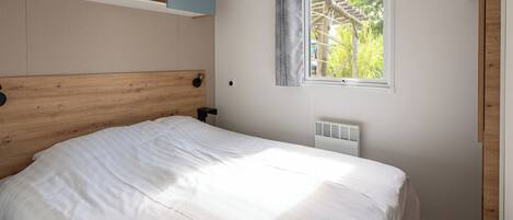 A bedroom with a wooden headboard, a white bedspread, a window with a view of greenery, and built-in shelving.