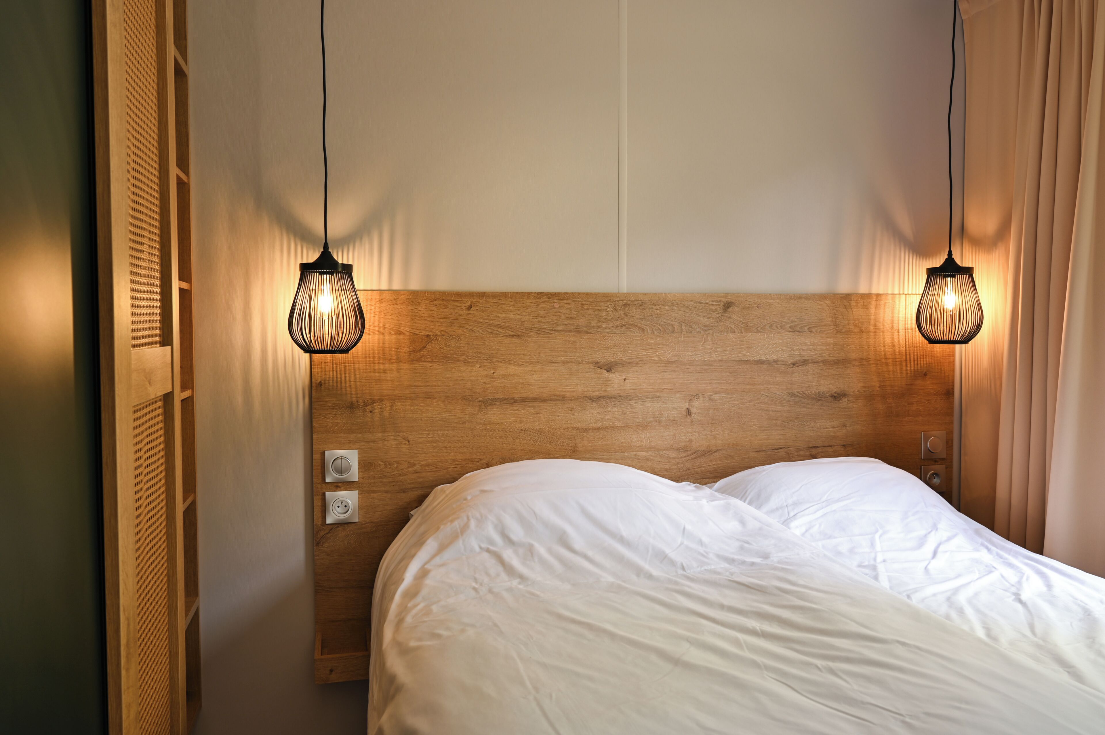 A neatly made bed with a wooden headboard, two pendant lights, and a power outlet on the wall.