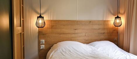A neatly made bed with a wooden headboard, two pendant lights, and a power outlet on the wall.