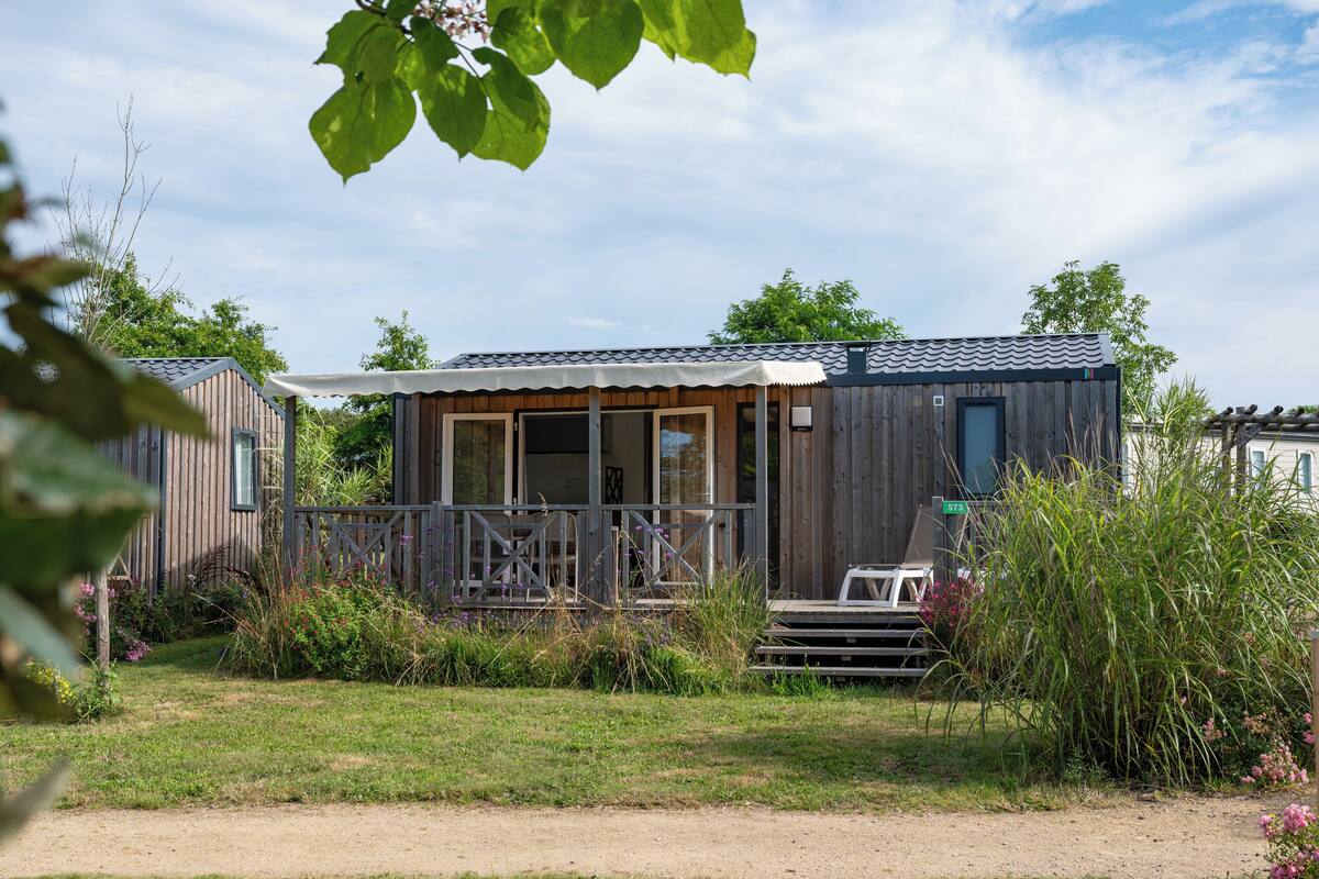 Eine Holzhütte mit Veranda, umgeben von Grünpflanzen und blauem Himmel.