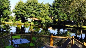 A deck with a table and chairs overlooking a calm lake, surrounded by lush greenery and trees. - RCN Camping La ferme du Latois (Coëx)