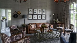 A living room with a brown leather sofa, patterned armchairs, a coffee table, and a chandelier. There are framed pictures on the wall and a window with curtains.