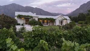 Une maison à deux étages, à l’extérieur blanc, entourée d’une végétation luxuriante et avec des montagnes en arrière-plan.