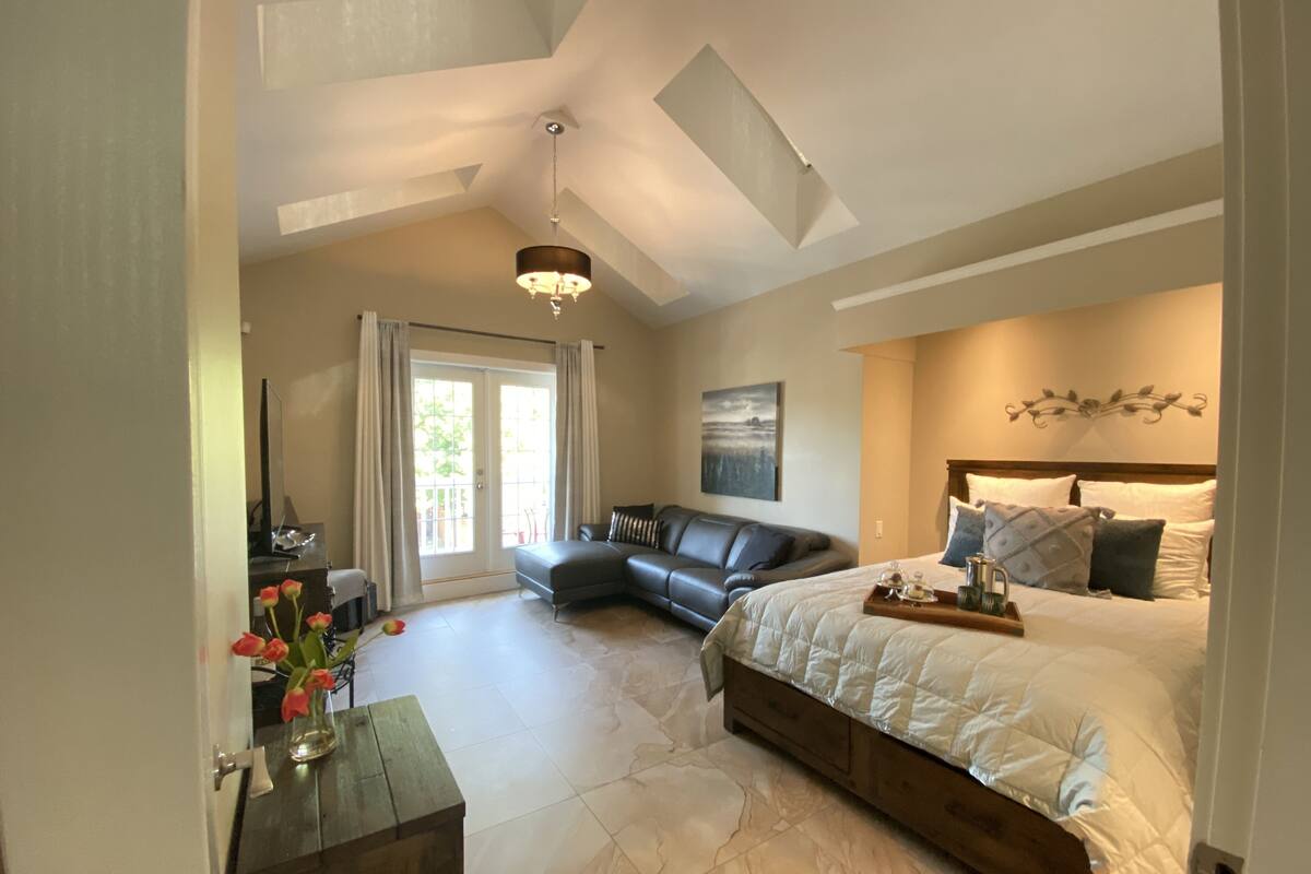 A bedroom with a large bed, a sofa, a desk with a vase of flowers, and a view of the outdoors.