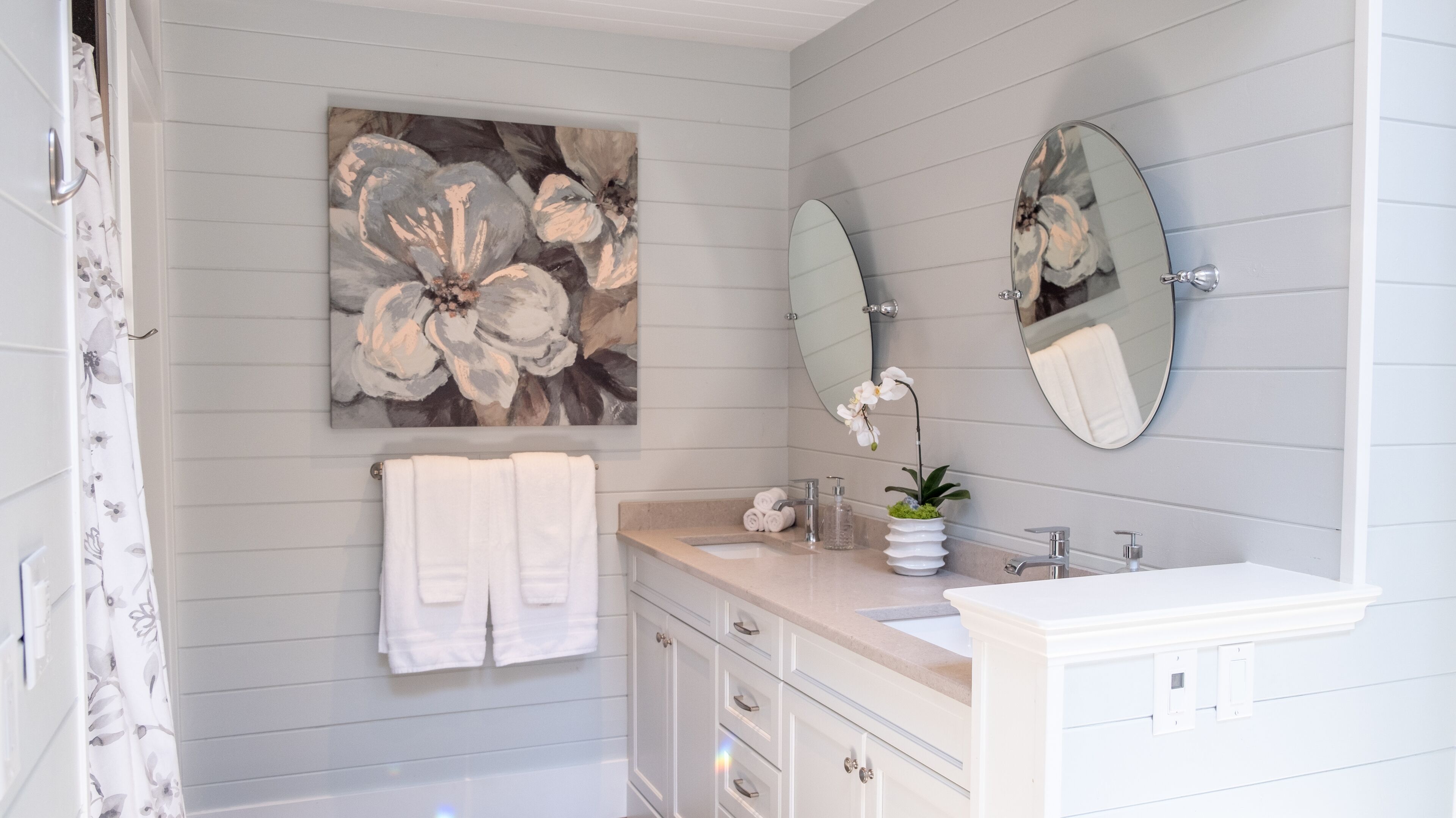 Une salle de bain avec un meuble-lavabo blanc, deux miroirs, un tableau représentant des fleurs et un rideau de douche.