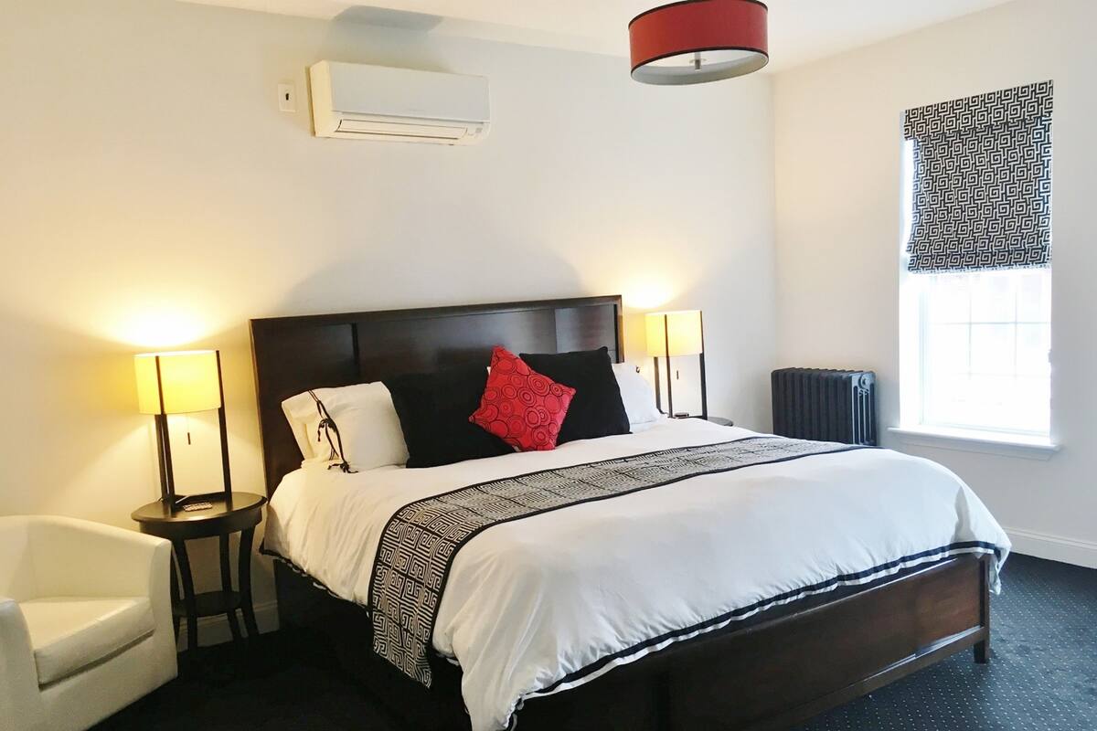 A hotel room with a large bed, two bedside lamps, a chair, a radiator, and a window with patterned curtains.
