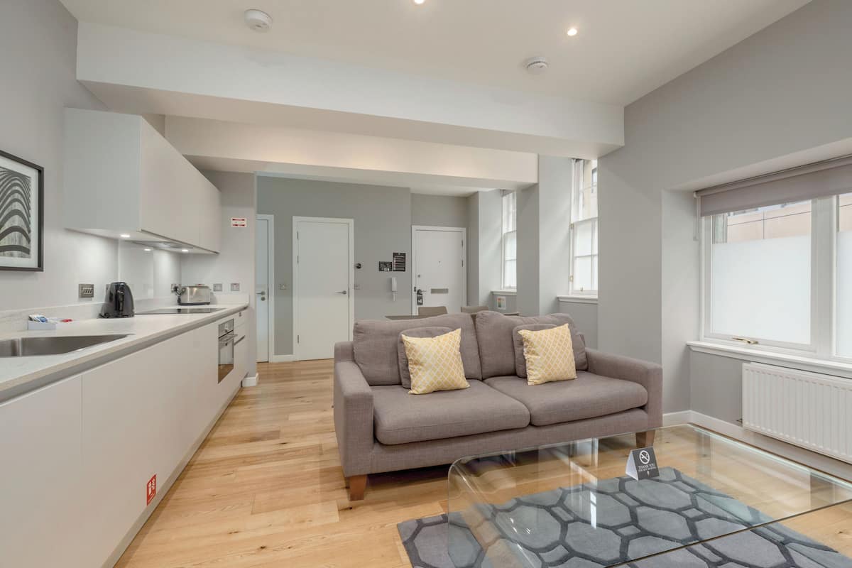 A modern living room with a sofa, a glass coffee table, and a kitchen area with a sink and appliances.