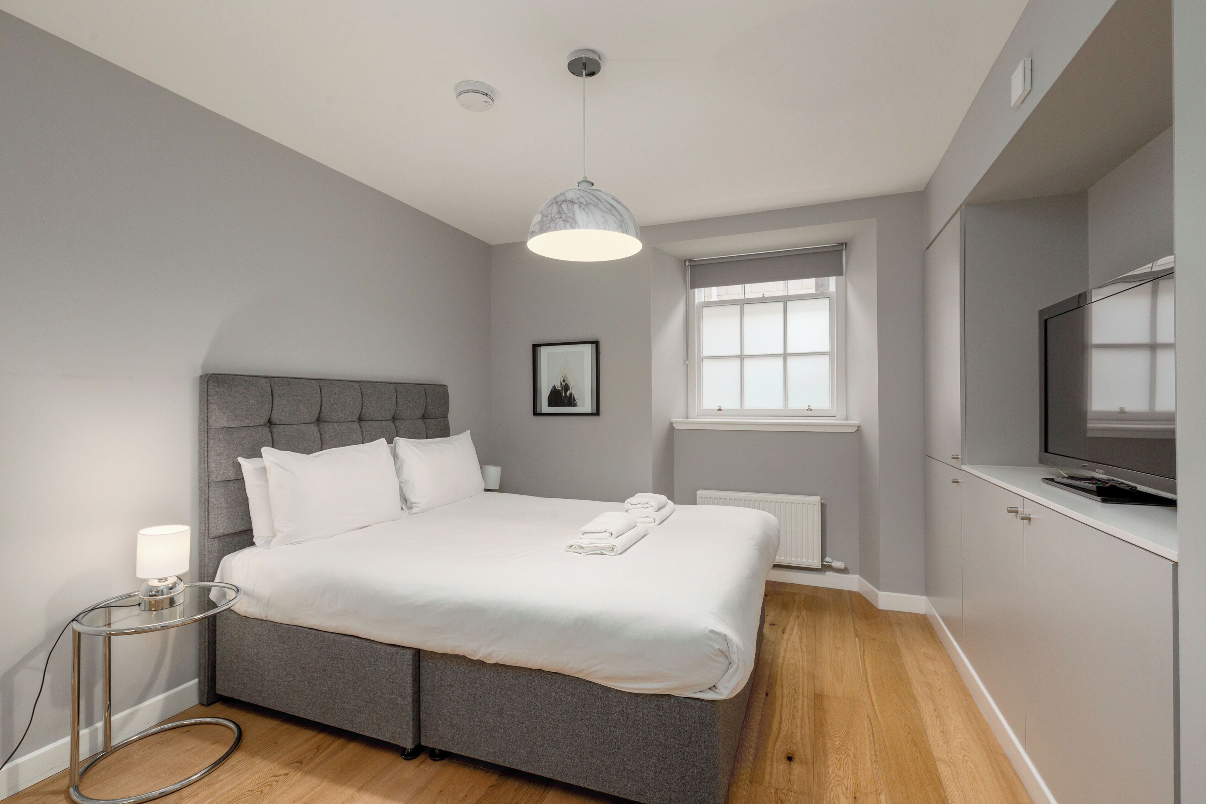 A modern bedroom with a grey upholstered bed, a bedside lamp, a framed picture on the wall, a window with blinds, and a TV on a cabinet.