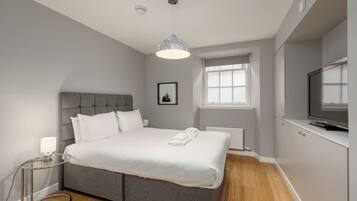 A modern bedroom with a grey upholstered bed, a bedside lamp, a framed picture on the wall, a window with blinds, and a TV on a cabinet.