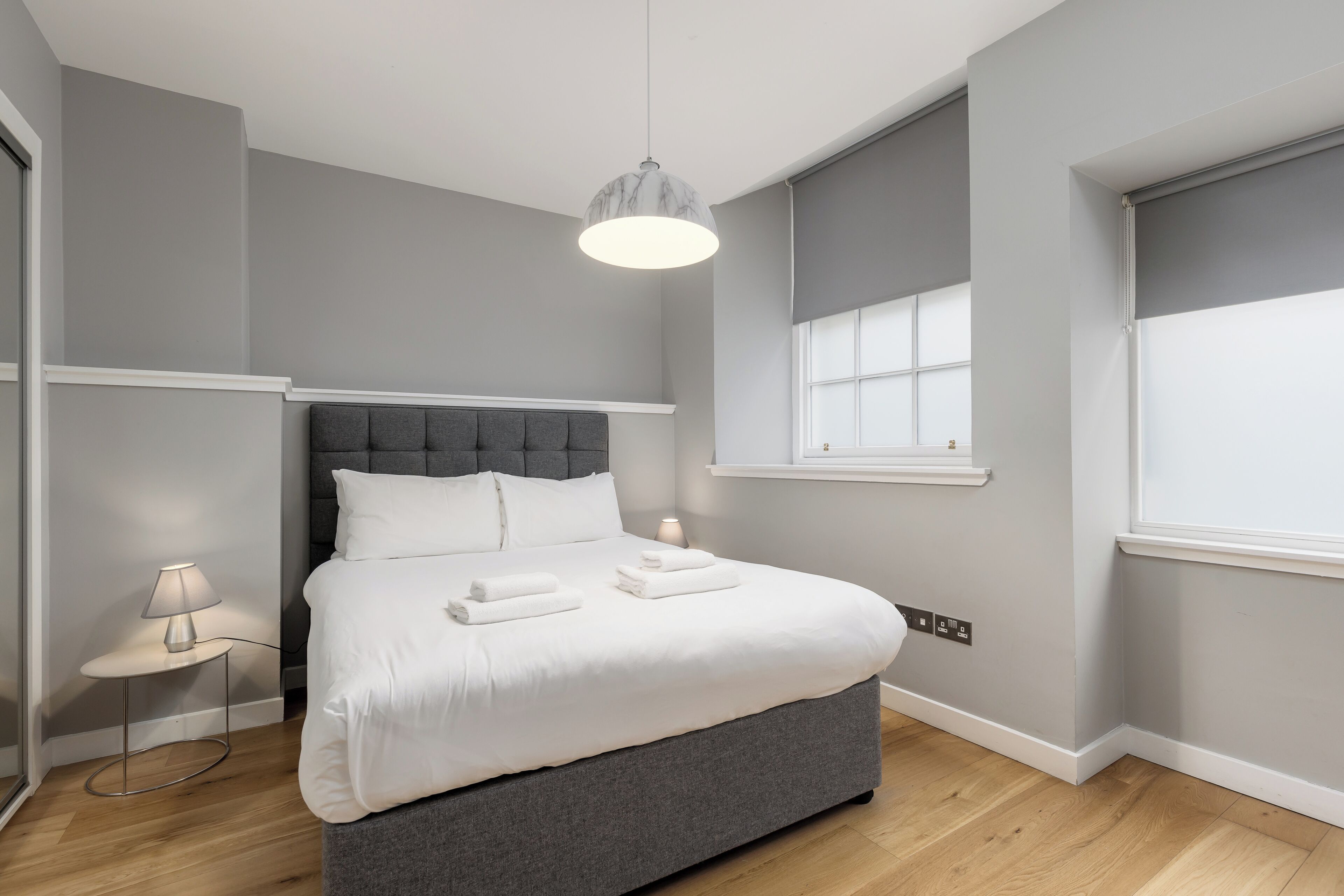 A modern bedroom with a grey upholstered bed, a bedside table with a lamp, and a window with blinds.