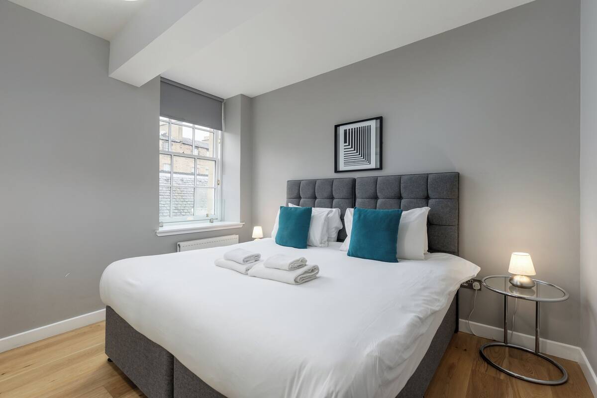 A modern bedroom with a large bed, grey walls, a window, and a bedside table with a lamp.