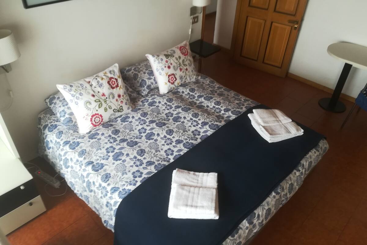 Un llit ben arreglat amb roba de llit i coixins de motius florals, una taula de nit amb una làmpada i una porta de fusta al fons.