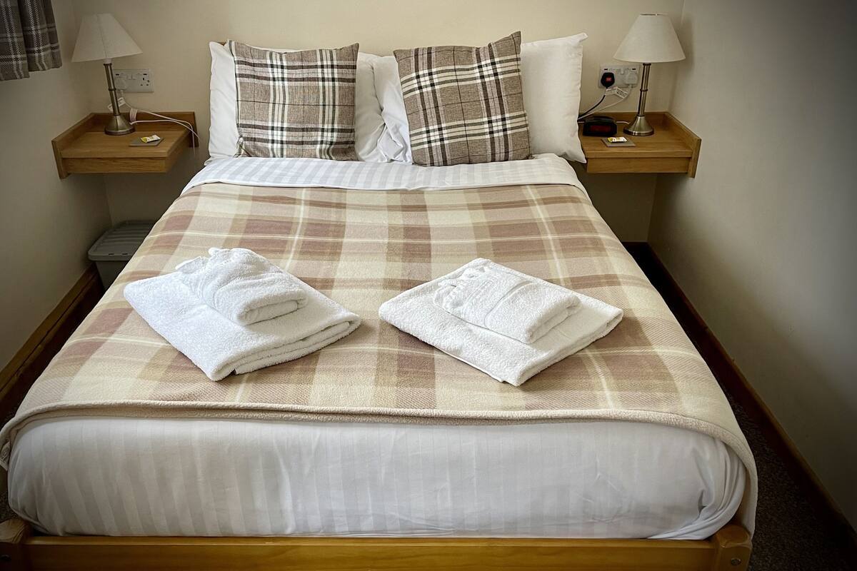 A neatly made bed with a plaid bedspread, two white towels, and two bedside lamps on wooden nightstands.