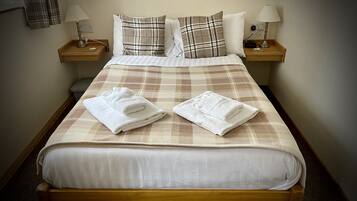 Una cama bien hecha con cubrecama escocés, dos toallas blancas y dos lámparas de noche sobre mesitas de madera.
