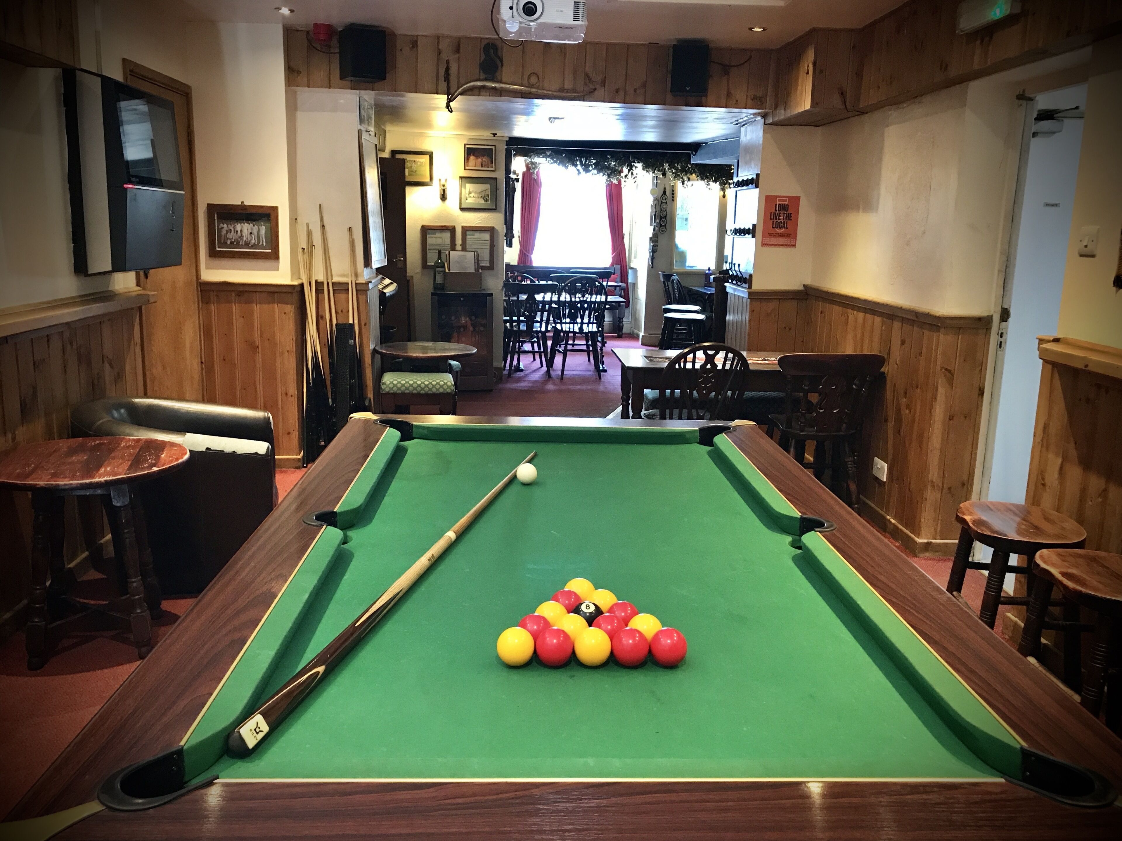 Una mesa de billar con bolas y tacos, paredes de madera y una zona de bar al fondo.