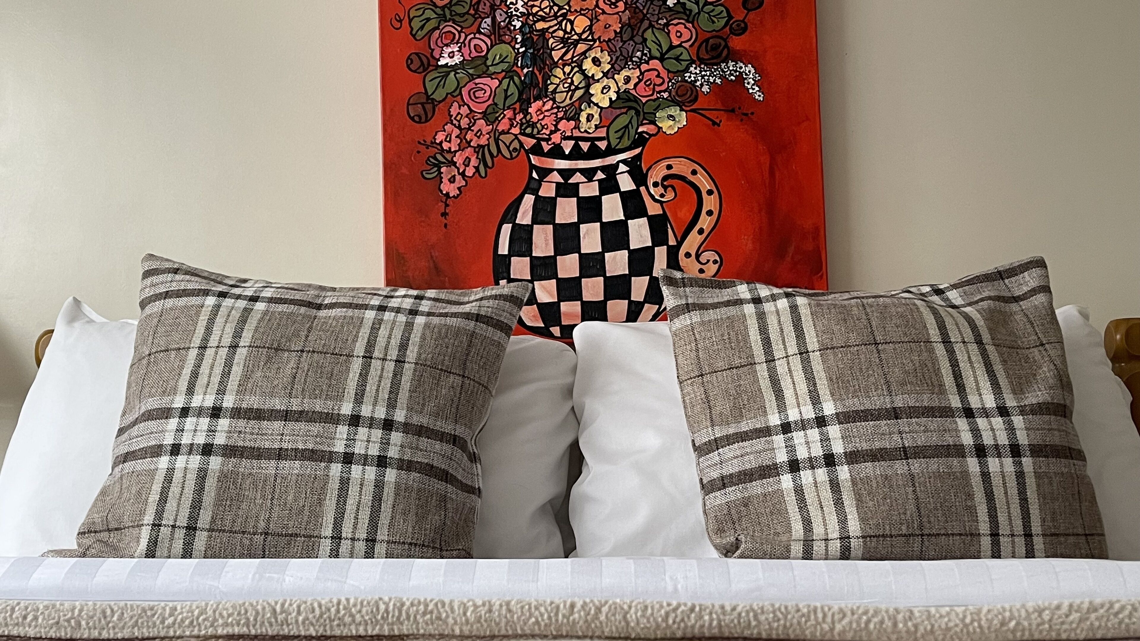 A bed with plaid pillows and a white pillow, a red floral painting on the wall, and a wooden headboard.