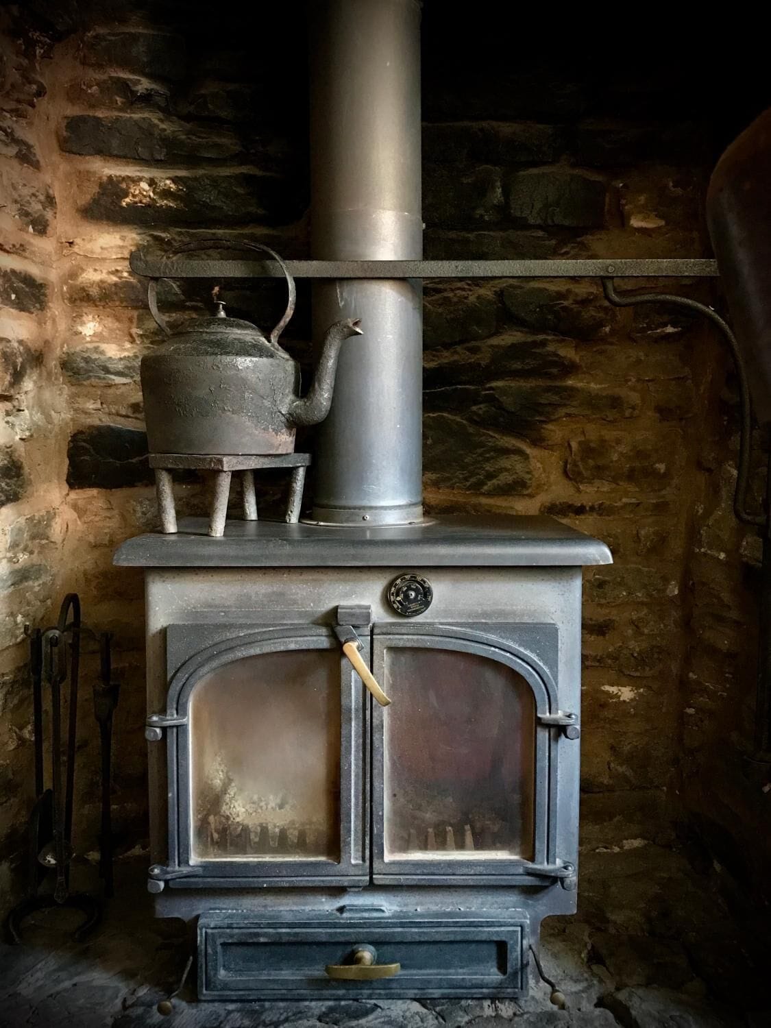 Una estufa de leña tradicional con una chimenea metálica, una tetera encima y un rústico muro de piedra de fondo.
