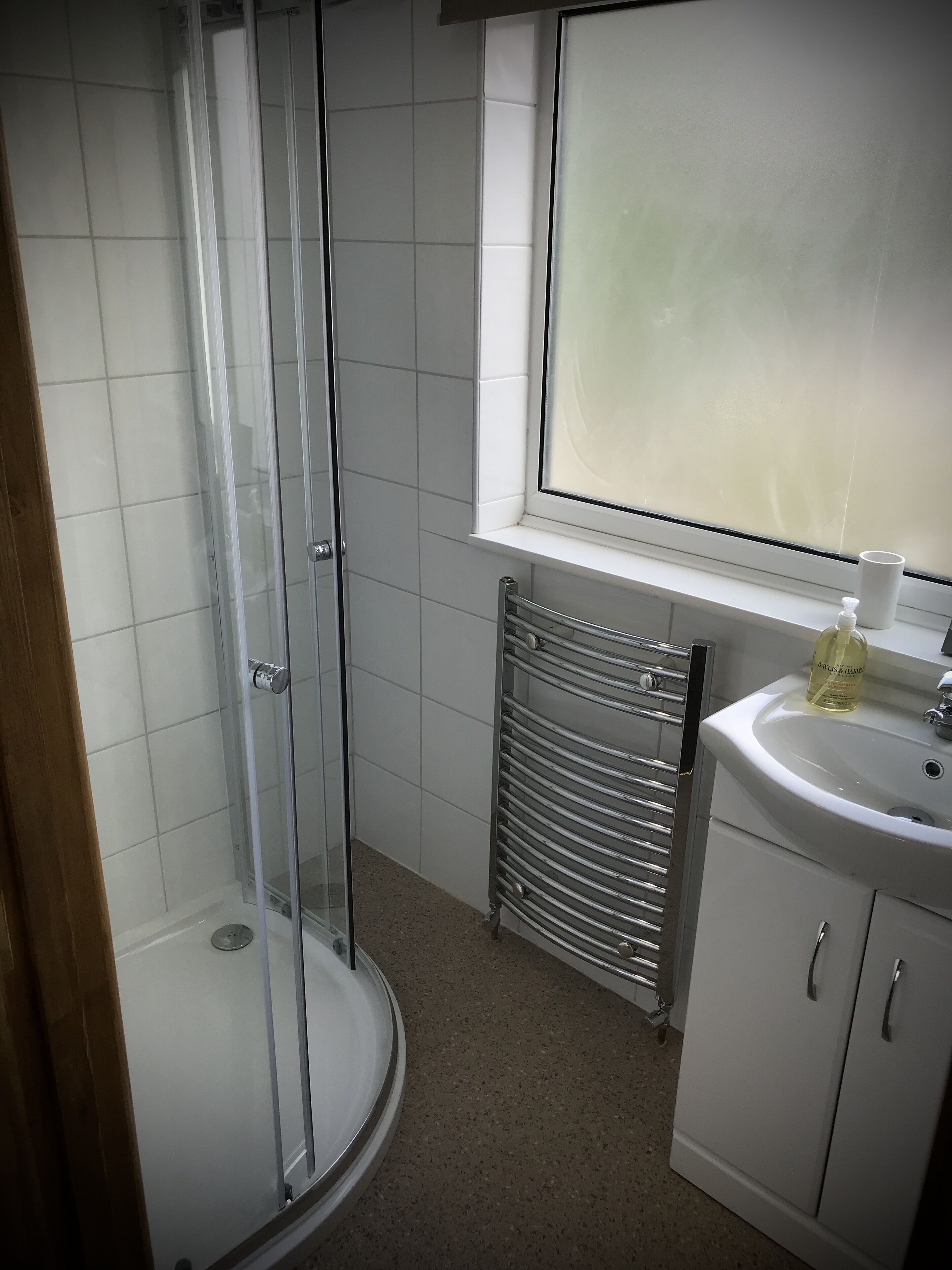 A bathroom with a glass-enclosed shower, a white sink with a bottle of liquid soap, and a window above the sink.