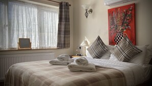 Una cama bien hecha con una manta y almohadones a cuadros, una mesita de noche con una lámpara, un cuadro en la pared y una ventana con cortinas.