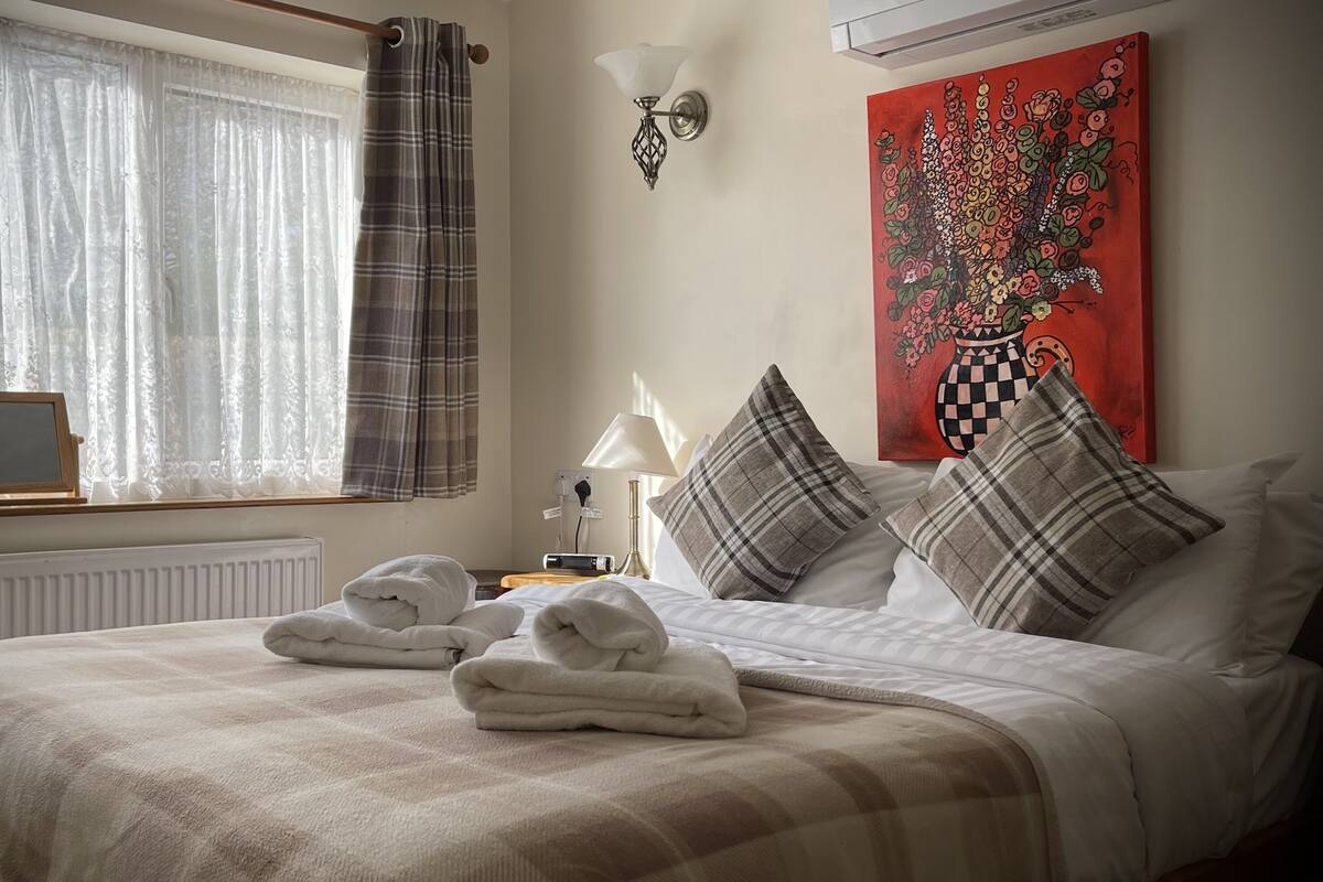 A neatly made bed with a plaid blanket and pillows, a bedside table with a lamp, a painting on the wall, and a window with curtains.