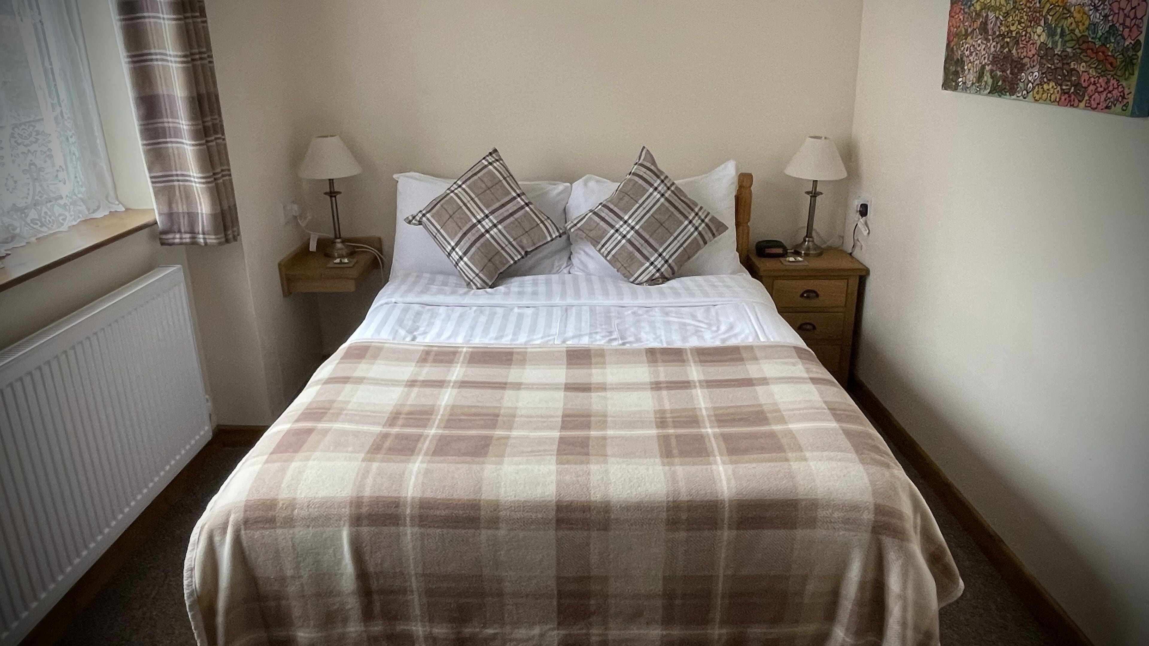 Una cama bien hecha con una colcha escocesa, dos almohadas a juego, dos mesitas de noche con lámparas y una ventana con cortinas.