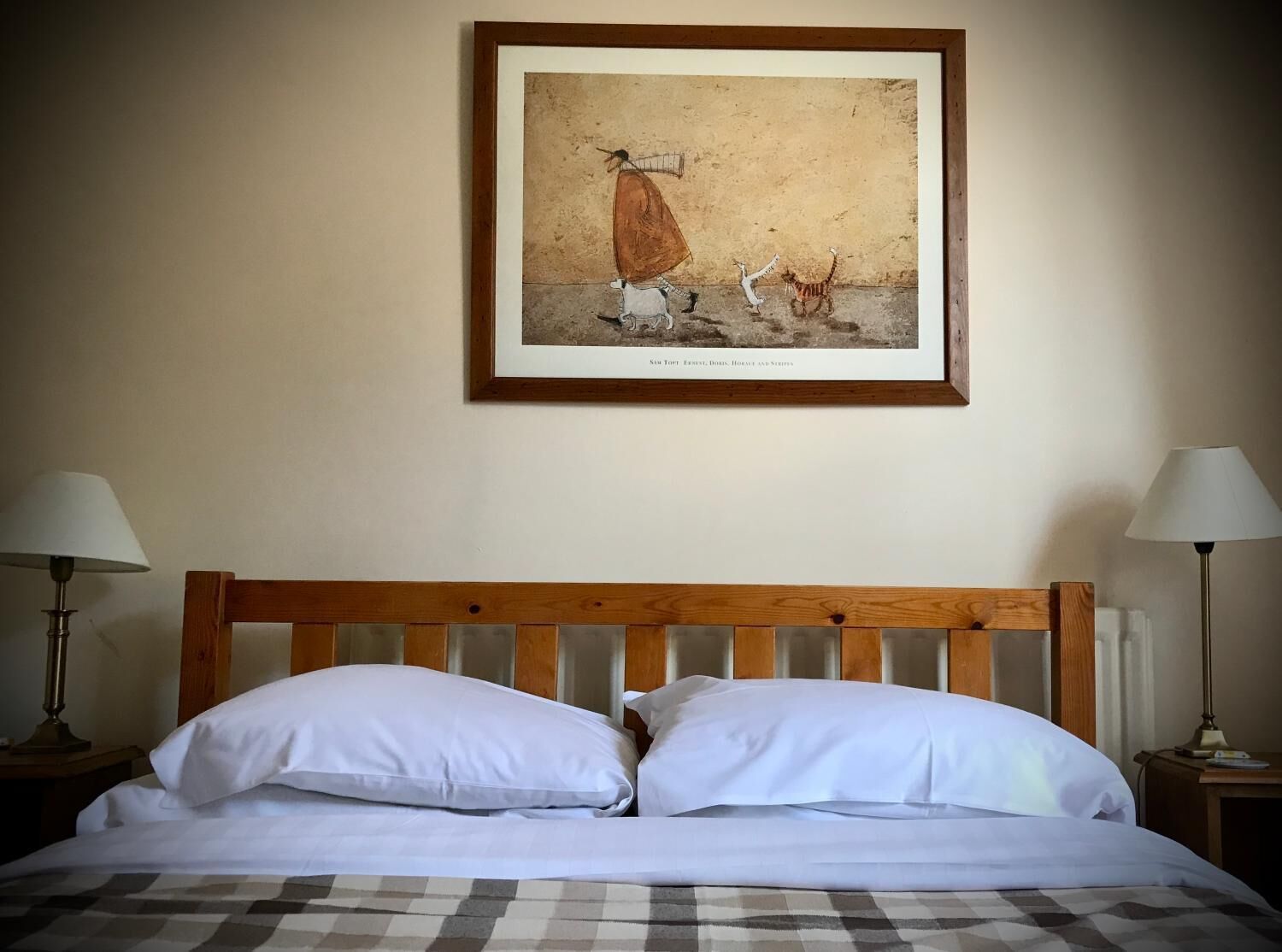 Un dormitorio con cabecera de madera, dos almohadas blancas, colcha a cuadros y un cuadro enmarcado de un caballo y un gato en la pared.