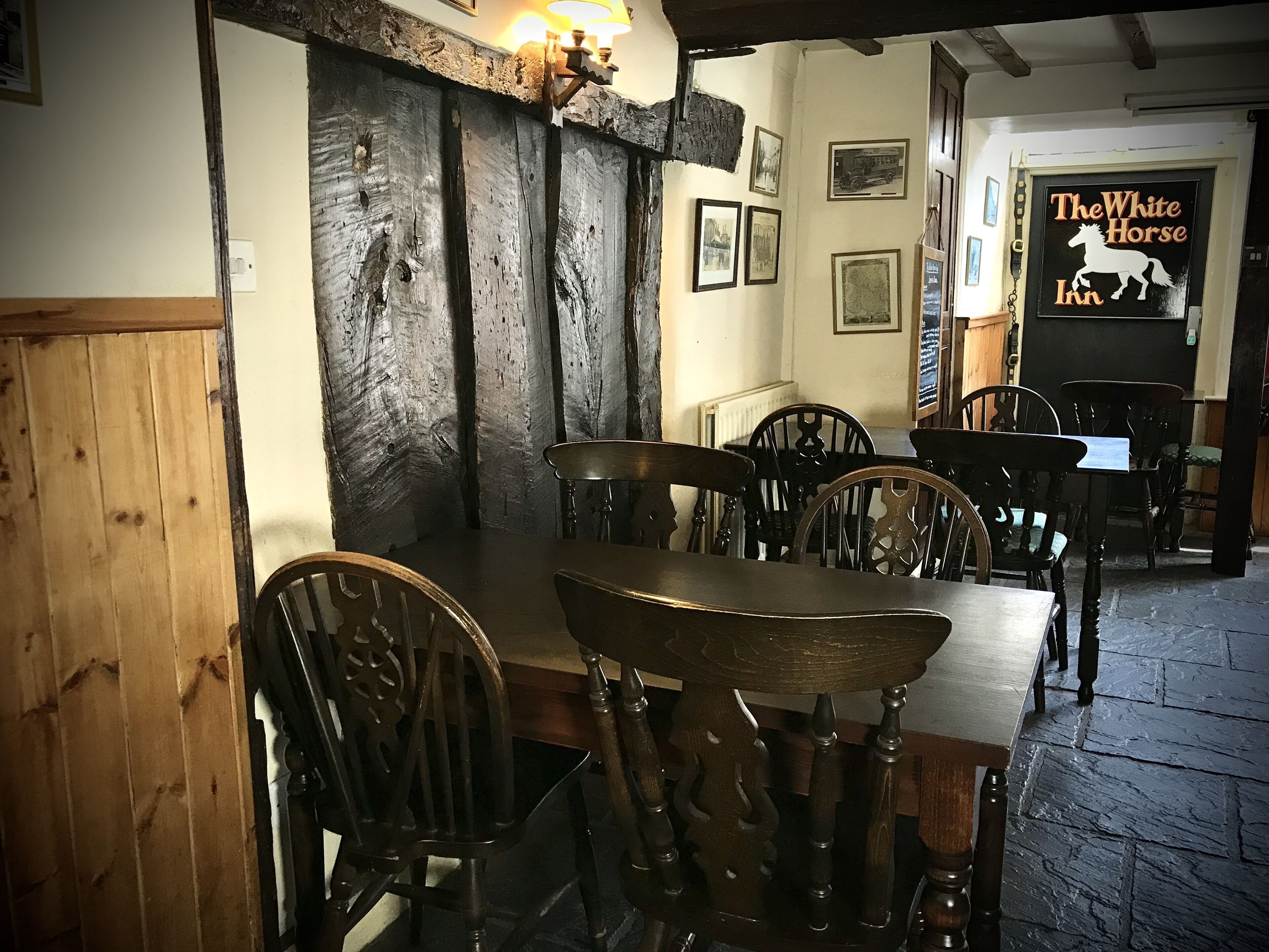 L’intérieur est rustique, avec des murs en bois et une longue table entourée de chaises. Des tableaux encadrés ornent les murs, et une enseigne indique « The White Horse Inn ».