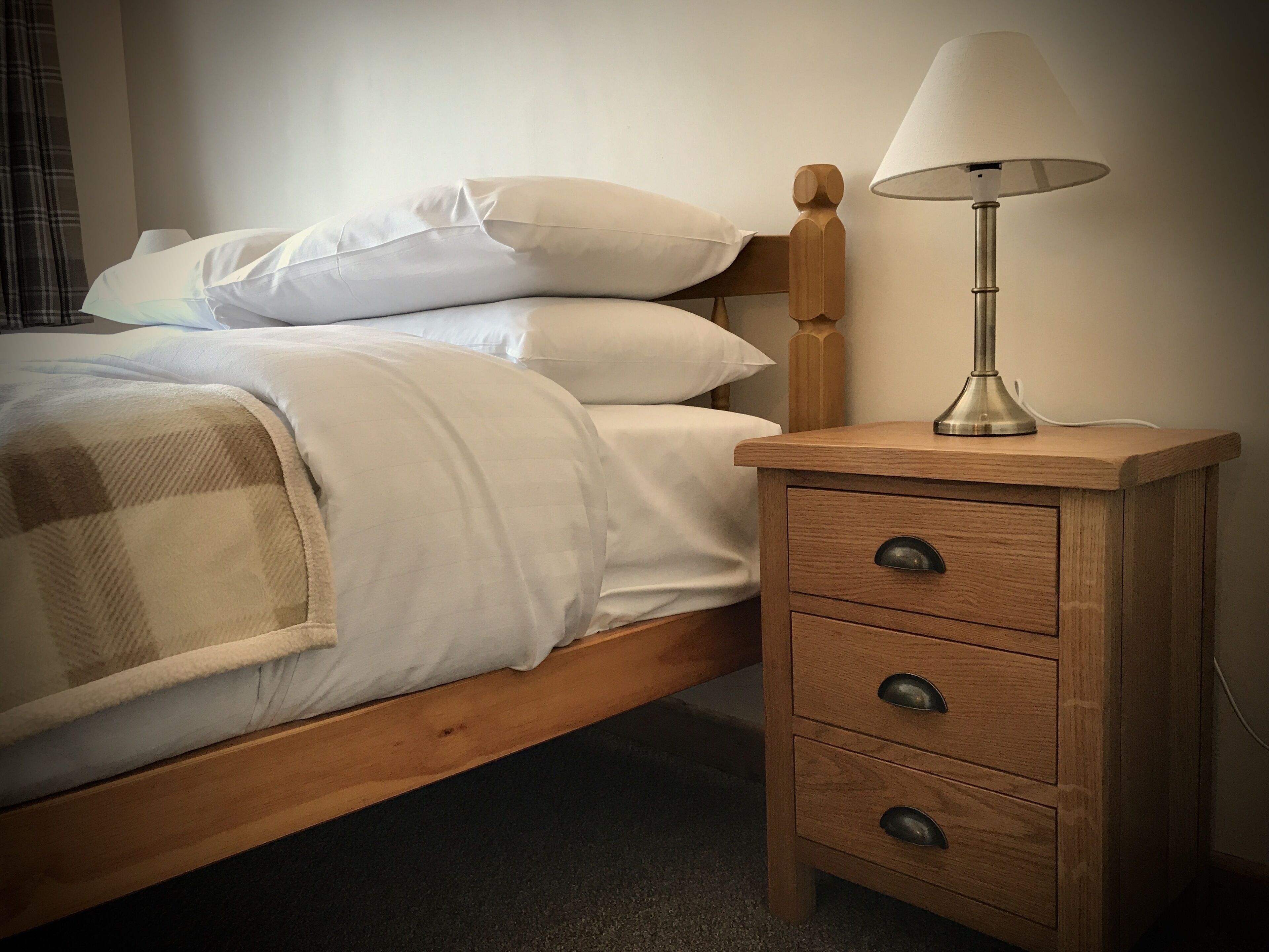 A bed with white linens and a wooden headboard, a bedside table with a lamp, and a checkered throw blanket.