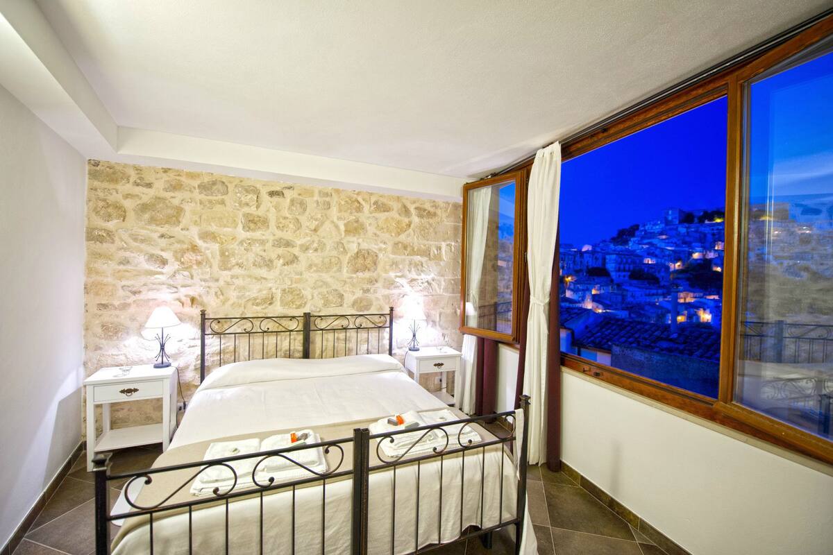 A bedroom with a stone wall, a large window with a view of a city at dusk, a bed with white linens, and two bedside tables with lamps.