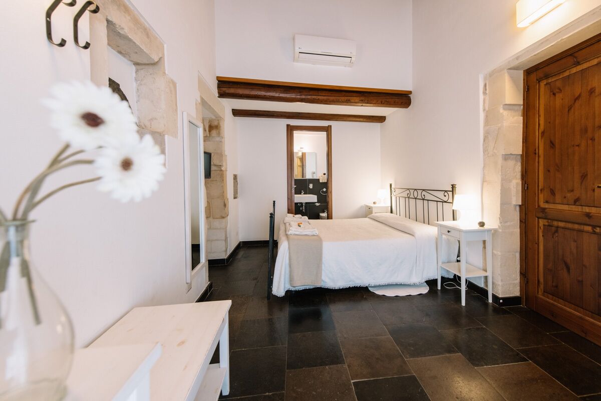 A bedroom with a bed, bedside table, and a mirror. There is a wooden door and a window.