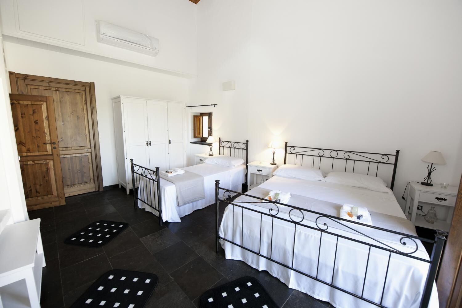 Une chambre d’hôtel avec deux lits, des murs blancs, une porte en bois et un sol carrelé.