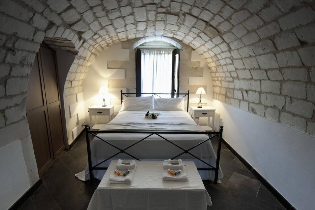 A bedroom with a stone arched ceiling, a bed with a white bedspread, two bedside tables with lamps, and a small table with towels and toiletries.