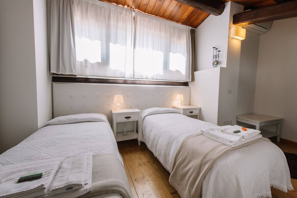 A bedroom with two single beds, a window with sheer curtains, bedside lamps, and a small table with a lamp.